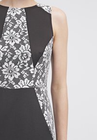 Zwarte jurk met een witte kanten bloemenoverlay op het lijfje, voorzien van een aansluitende snit en zonder mouwen. Soepele textuur en geometrische vorm.