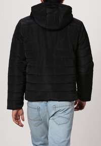 Sort pufferjakke med hætte, der har lodret quiltede syninger og en let cropped pasform, set fra bagsiden.