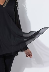 Žena v volné černé blůze s průsvitnými rukávy a zdobeným výstřihem, s jednou paží nataženou, vrhající stín na bílou zeď.