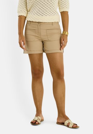 Femme portant un short beige, un haut en maille crème, des bracelets dorés, des bagues et des sandales beige à bout ouvert, debout sur un fond uni.