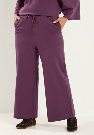 Persoon draagt losse paarse broek met elastische taille en trekkoord, handen in de zakken, gecombineerd met zwarte en bruine sneakers met patroon.