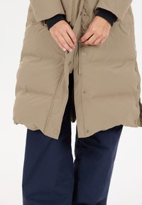 Beige pufferjakke med lynlås og trykknaplukning, der har en quiltet tekstur. Sammen med marineblå bukser, hvor hænderne er placeret nær jakken.