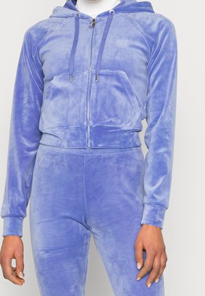 Woman wearing a soft, light blue velour zip-up hoodie and matching pants, with hands relaxed at sides.