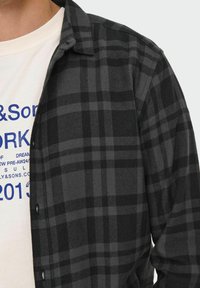 Zwart en grijs geruite flanellen overhemd met knoopsluiting en kraag, gedragen over een wit T-shirt met blauwe tekst en afbeeldingen.