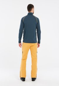 Tamsiai mėlynas vyriškas sportinis švarkas su aukšta apykakle ir geltonomis siūlėmis, derinamas su geltonomis kelnėmis, turinčiomis šoninius užtrauktukus, ir juodais batais.