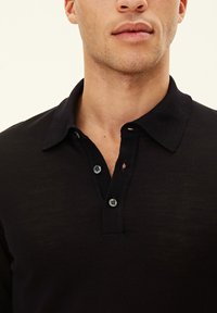 Svart polo t-shirt med krage, som har två knappar och ett texturerat tygmönster. Närbild som betonar sömmar och knapppdetaljer.