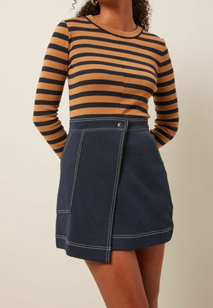 Femme portant un haut à manches longues rayé marron et noir et une jupe bleu marine avec des coutures blanches et un détail de bouton-pression à l'avant.