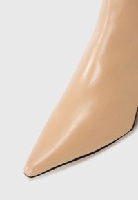 Chaussure à talon haut en cuir beige avec un bout pointu et une texture lisse. Semelle noire visible à la base, mettant en valeur un design élégant.