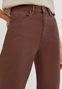 Jeans en denim marron avec une finition texturée, taille haute, poches avant et détails en métal doré. Vue en gros plan mettant en valeur les détails des coutures.