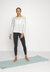 Light gray long-sleeve top with a round neckline, paired with black leggings. Model stands on a textured light blue yoga mat.