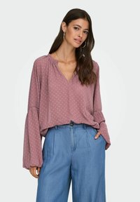 Kvinde med langt mørkt hår iført en løs mauve bluse med lange ærmer og lyseblå højtaljede bukser med brede ben, stående mod en hvid baggrund.