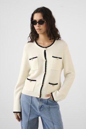 Ženska z rjavimi sončnimi očali, kremasto teksturiranim cardiganom s črnim robom in zlatimi gumbi, oblečenimi z ohlapnimi svetlo modrimi kavbojkami, z roko v žepu.