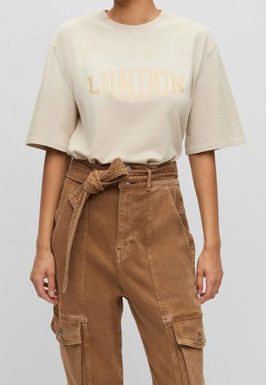 Osoba nosząca beżowy t-shirt "LONDON" włożony w brązowe spodnie z wysokim stanem, z przewiązanym paskiem i dużymi zapinanymi na guziki kieszeniami.
