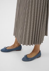 Denim Ballerinas haben eine runde Zehenpartie, eine Schleifenakzent und einen hellbraunen Holzabsatz. Sie werden mit einem plissierten, karierten Rock getragen.