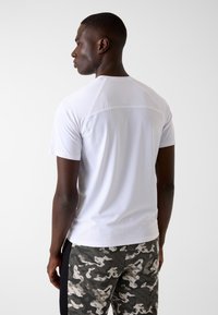 Weißes Sportshirt mit kurzen Ärmeln und strukturiertem Mesh-Rücken, kombiniert mit Hosen im Camouflage-Muster, die schwarze Akzente und hellere Farbtöne aufweisen.