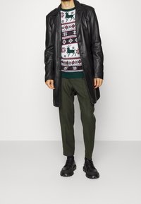 Schwarzer Leder-Trenchcoat über einem gemusterten Pullover mit Rentieren und geometrischen Designs, kombiniert mit olivgrünen Hosen und schwarzen Schuhen.