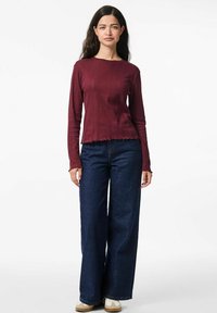 Bordo ilga rankovė viršūnė su raguotomis tekstūromis ir skeltukais apačioje, su derančiais aukštu juosmeniu tamsiai mėlynomis plačiomis džinsų kelnėmis ir smėlio spalvos sportiniais bateliais.