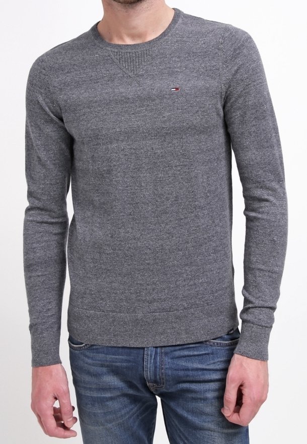 Homme portant un pull gris ajusté à col rond avec un petit logo rouge, blanc et bleu sur la poitrine et un jean bleu.