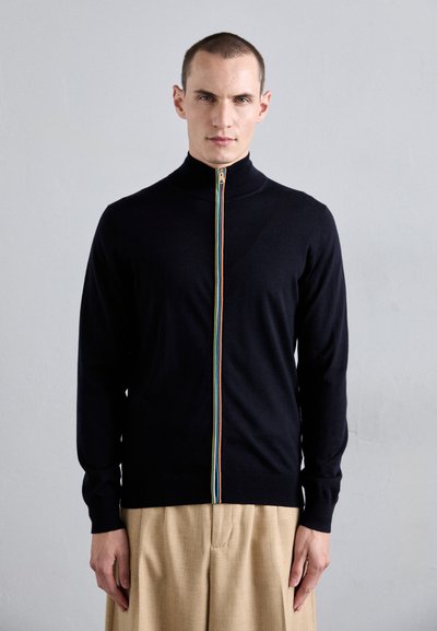 Schwarzer Reißverschluss-Pullover mit hohem Kragen, ausgestattet mit einem multicolor Reißverschluss. Das Modell trägt ihn zu heller, weit geschnittener Hose.