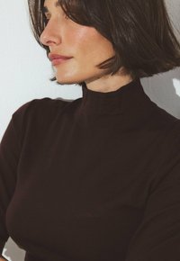 Brown ribbed turtleneck sweater with a fitted design, soft texture, and a high neckline, worn by a person posed against a neutral backdrop.
