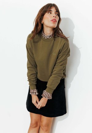 Sweat-shirt vert olive avec poignets côtelés, superposé sur une chemise à carreaux. Le modèle porte une jupe en velours côtelé noire, les mains jointes à la taille.