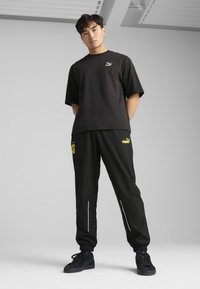 Camiseta de algodón negra con el logotipo de Puma, combinada con pantalones de chándal negros con acentos amarillos y detalles reflectantes, y zapatillas negras.