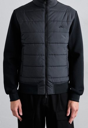 Veste matelassée noire à manches longues avec fermeture éclair devant, portée par une personne visible des épaules aux hanches sur un fond uni.