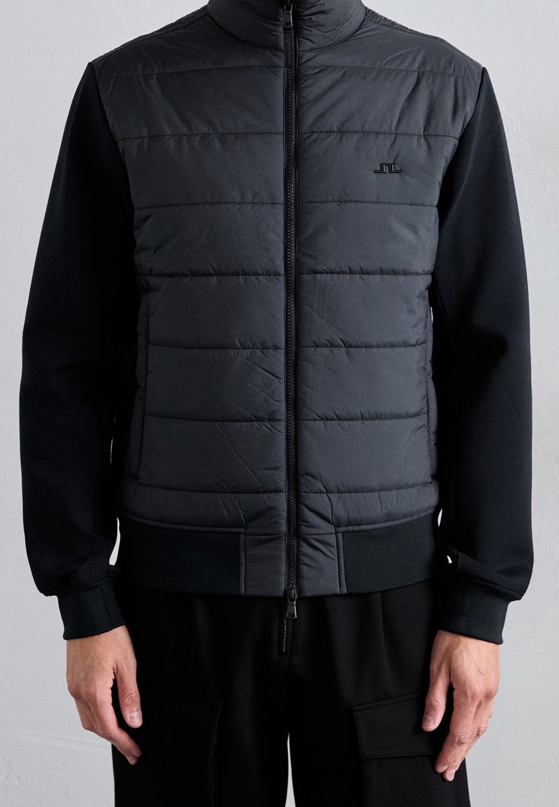 Veste matelassée noire à manches longues avec fermeture éclair devant, portée par une personne visible des épaules aux hanches sur un fond uni.