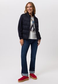 Navy puffer jacket with a ribbed collar, worn over a light graphic t-shirt. Paired with dark denim jeans and red platform sneakers.