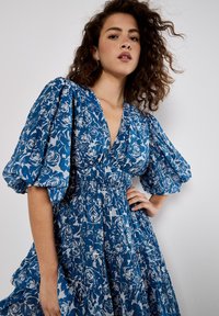 Blau-weißes Kleid mit Floralprint, Puffärmeln, tiefem V-Ausschnitt und gestuftem Saum. Das Material wirkt leicht und hat eine weiche Textur.
