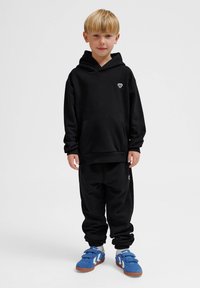 Schwarzer Kapuzenpullover mit Kängurutasche, kombiniert mit schwarzen Jogginghosen. Blaue Sneaker mit Klettverschlüssen runden das Outfit ab.