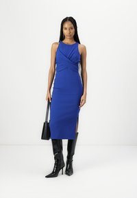 Rochie midi bleu fără mâneci, cu detalii pliate la bust și fanta pe lateral, asortată cu cizme negre cu toc stilettos până la genunchi și o geantă de umăr.