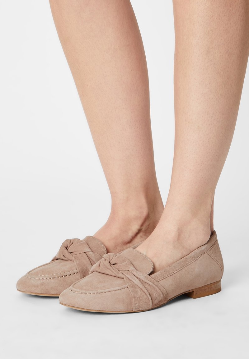 Mocassins en daim beige avec un détail noué à l'avant, semelle plate et accents cousus. Présentent une texture douce et un design à bout arrondi.