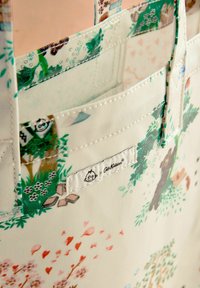 Sac fourre-tout réutilisable en tissu crème avec des motifs naturels colorés, incluant des arbres et des animaux. Dispose de deux poignées robustes et d'une étiquette de marque.