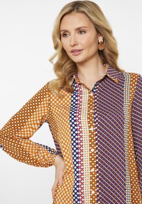 Multifarvet bluse med geometriske mønstre i orange, blå og lilla. Knappet front med lange ærmer og krave.