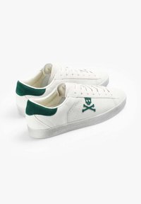 Zapatillas de ante blancas con acentos de ante verde en el talón, que presentan un logo de calavera y huesos cruzados, suelas de goma texturizadas y cordones planos.