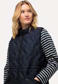 Marineblaues Steppweste mit quilteter Oberfläche und hohem Kragen, kombiniert mit einem gestreiften Langarmoberteil. Mit einem Frontreißverschluss und zwei Taschen ausgestattet.