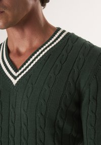 Zelený pletený svetr s kabelovým vzorem, designem do V a bílými/černými pruhovanými žebrovanými detaily na límci.