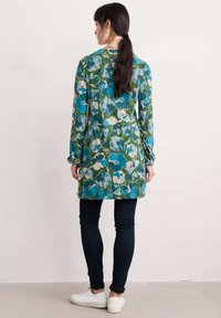 Túnica floral en verde, azul y blanco, con mangas largas y corte recto. Combinada con pantalones oscuros y zapatillas blancas.