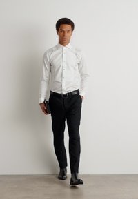 Fehér gombos ing, fekete slim-fit nadrág, fekete öv, fekete clutch, fényes fekete cipők, sima háttér előtt áll.