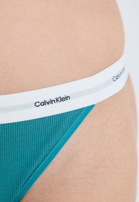 Tyrkis rillede bikini bund med en hvid elastisk talje med sort "Calvin Klein" logo. Glat tekstur og minimalistisk design.