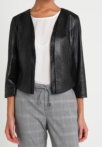 Femme portant une veste courte noire ouverte sur le devant, un haut blanc à col rond et un pantalon gris à carreaux avec cordon de serrage, sur un fond uni.