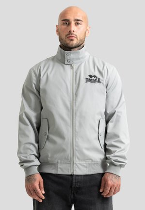 Hombre con tatuajes que lleva una chaqueta Lonsdale London gris claro con cremallera contra un fondo gris liso.
