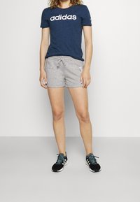 Person som bär en marinblå Adidas T-shirt, ljusgrå shorts med snörning och svarta Adidas sneakers med vita ränder, står på ett grått golv.