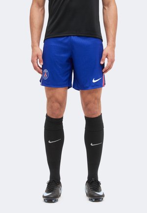 Egy személy lábai, aki kék Paris Saint-Germain focishortot, fekete Nike zoknit, fekete stoplis cipőt és fekete inget visel.