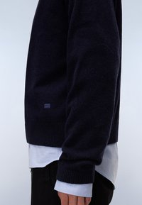 Dunkelblauer Pullover aus Strick mit gerippten Bündchen und Saum, der ein dezentes Logo-Detail an der unteren Seite aufweist. Über einem hellblauen Hemd getragen.