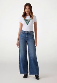 White t-shirt with green and leopard print triangle design, paired with high-waisted, wide-leg blue jeans, and black ankle boots.