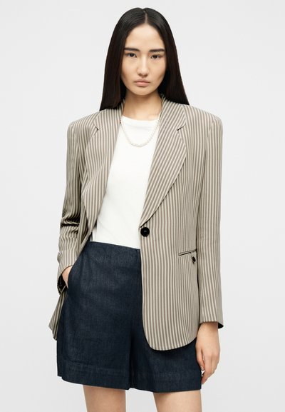 Γυναίκα με μακριά σκούρα μαλλιά που φοράει μπεζ ριγέ σακάκι, λευκή μπλούζα, κολιέ με πέρλες και σκούρα τζιν σορτς, πόζα δίπλα σε απλό φόντο.