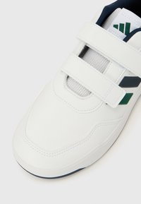 Balta sportinė avalynė su lygiu odiniu viršumi, akių tinklo detalėmis, dviem lipniais užsegimais ir mėlynais bei žaliais dizaino elementais.