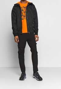 Sort letvægtsjakke med hætte over en orange grafisk t-shirt, parret med sorte cargo-bukser og sorte vandresko.